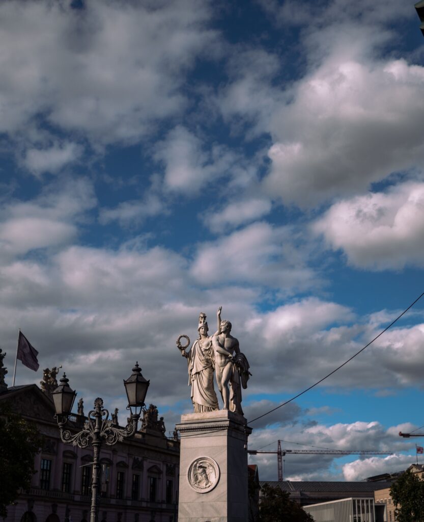 A classical sculpture atop a pedestal with a background of textured clouds and blue sky.