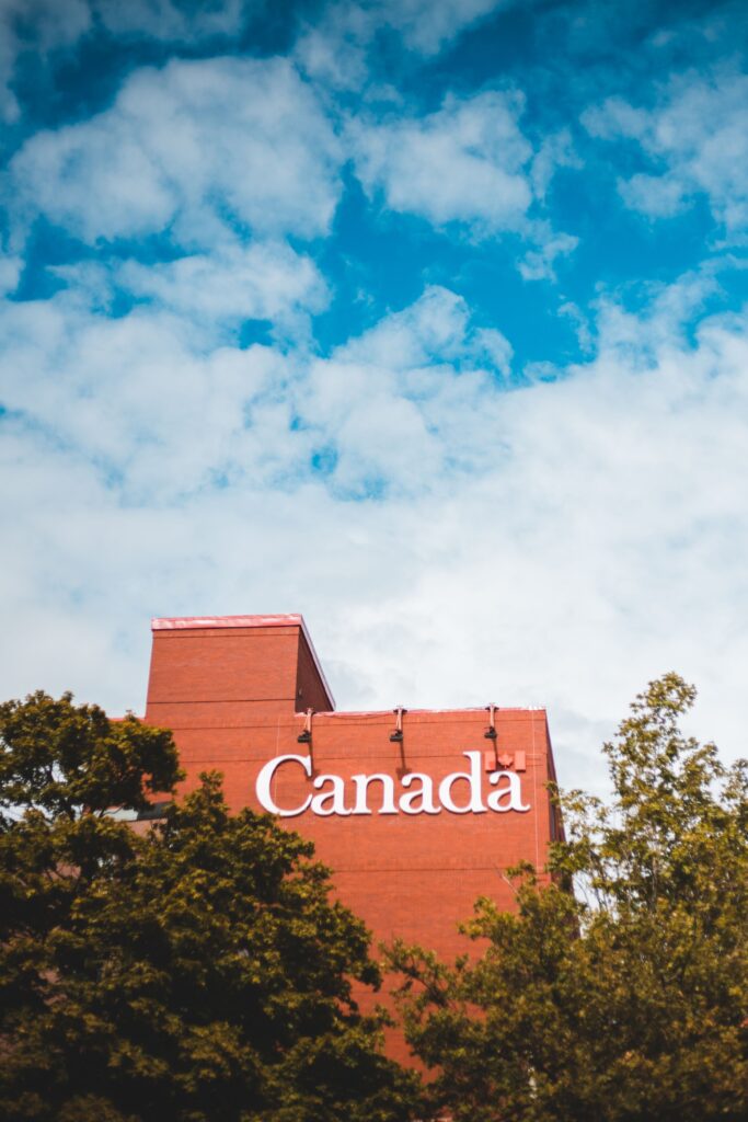 Red brick government building marked 'Canada' with trees in foreground and blue sky above.